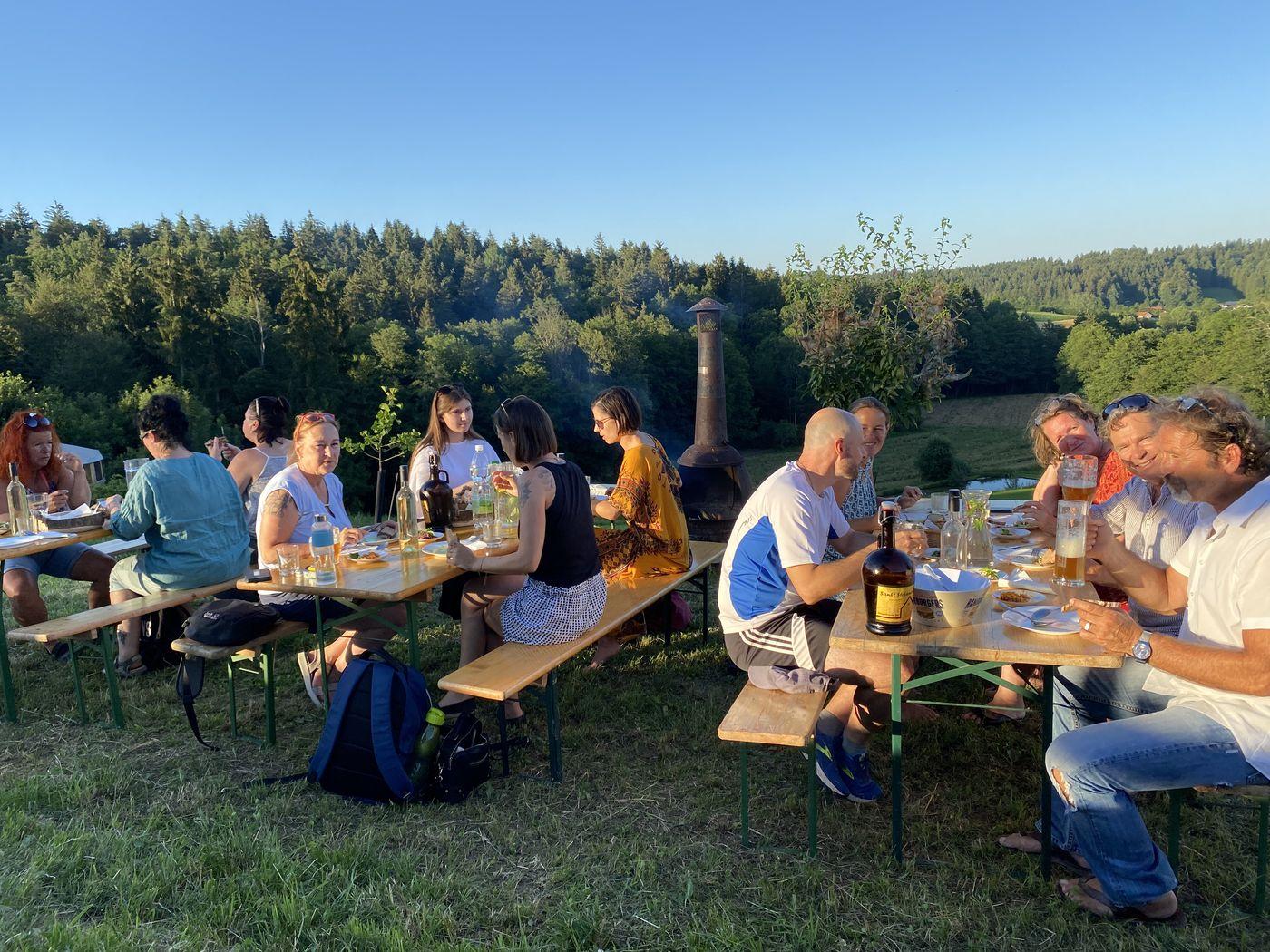 Gruppe sitzt in der Abendsonne an langen Holztischen im Freien mit Wald und Hügeln im Hintergrund
