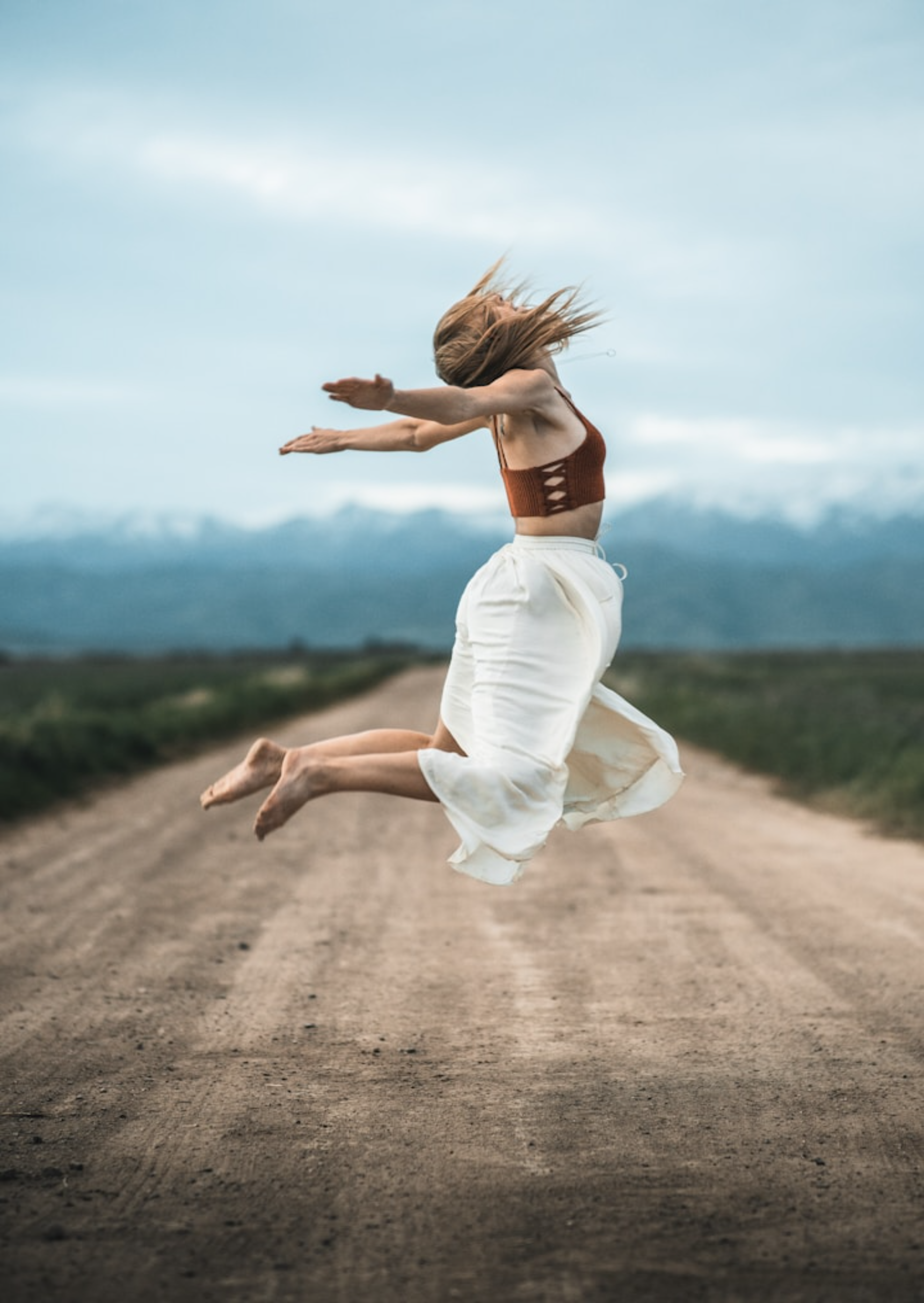 A blonde, barefoot woman leaping joyfully, a wide dirt road beneath her and blue mountains in the background,symbolising freedom