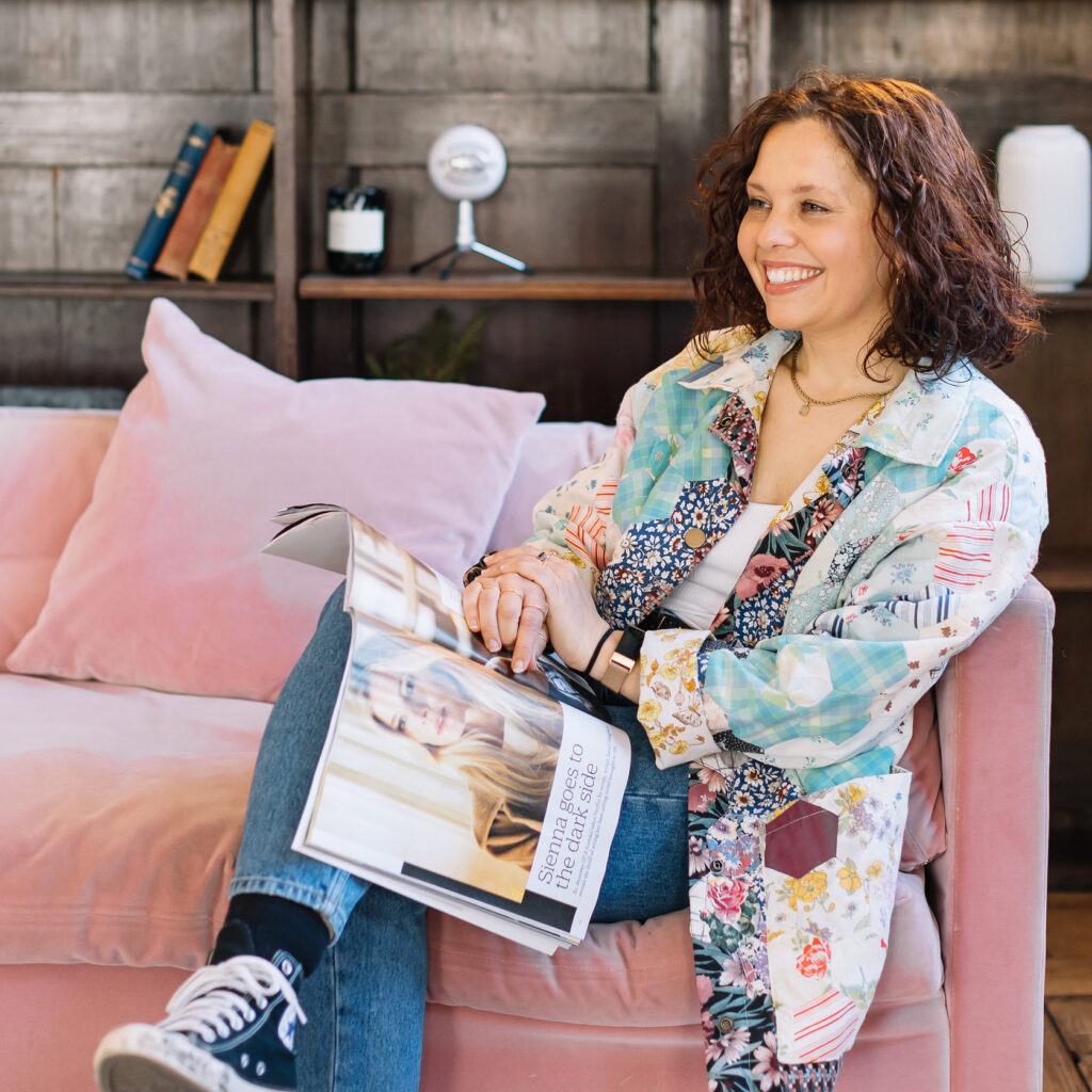 Elizabeth Stiles smiling on a pink sofa, holding an open magazine with the headline 'Sienna goes to the dark side', wearing a patchwork…