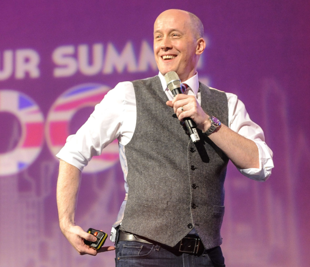Chris Ducker smiling on stage at a summit, holding a microphone and a presentation clicker, wearing a grey vest over a white shirt.