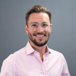 Levi, smiling professional wearing clear-frame glasses and a pink button-up shirt, headshot photo