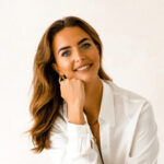 Hannah, a smiling woman with long brown hair wearing a white shirt, rests her chin on her hand in a professional headshot.