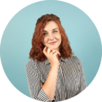 Smiling woman with curly red hair resting her chin on her hand, wearing a black and white striped shirt against a light blue background.
