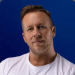 Headshot of Jason, a man with short brown hair and a beard wearing a white t-shirt against a blue background